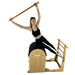 A woman performs a pilates exercise on a wooden reformer machine, holding a wooden bar overhead against a solid white background.