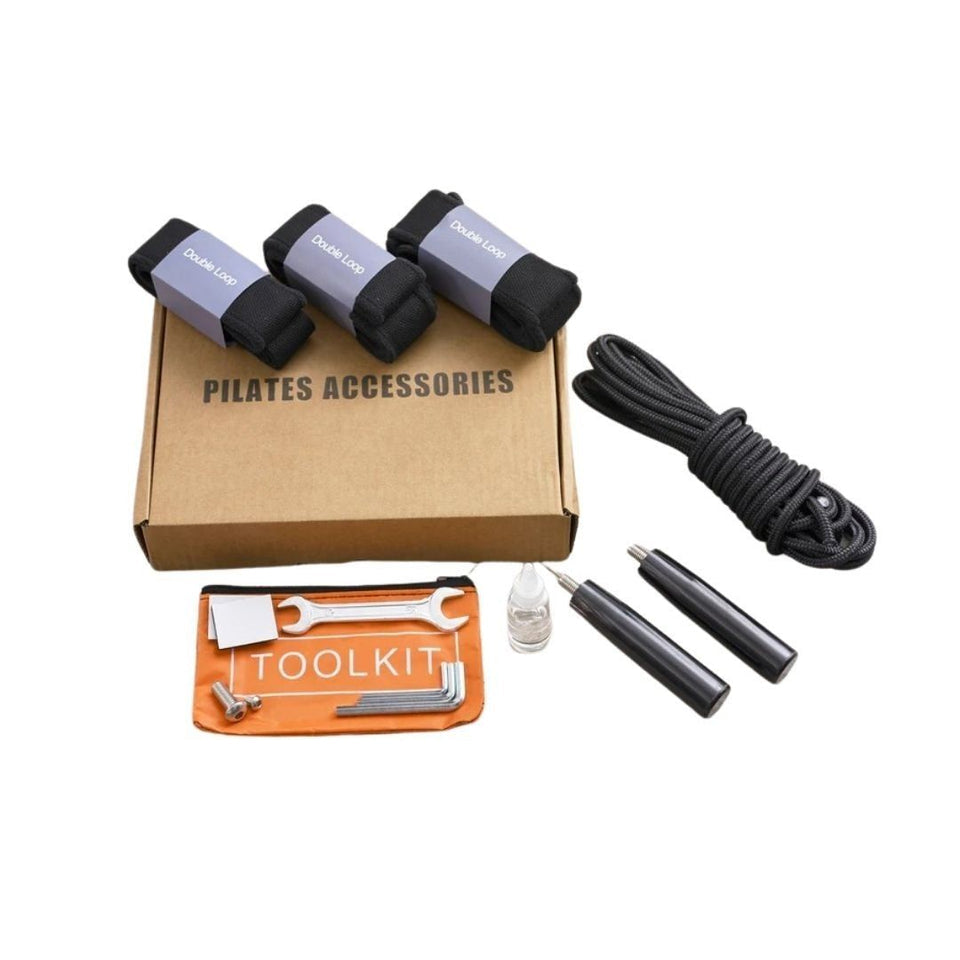 Pilates accessories kit including three black and gray fabric rolls on a brown box, a black coiled strap, two black handles, a small clear bottle, and an orange tool pouch with metal tools, labeled 'PILATES ACCESSORIES' and 'TOOLKIT'.