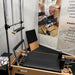 A wooden Pilates reformer machine with black padded straps and handles, shown indoors with an attached metal spring bar frame. A large printed poster and instruction panel are visible behind it.