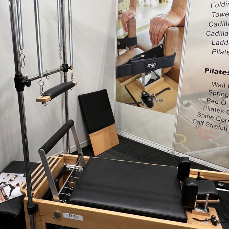 A wooden Pilates reformer machine with black padded straps and handles, shown indoors with an attached metal spring bar frame. A large printed poster and instruction panel are visible behind it.